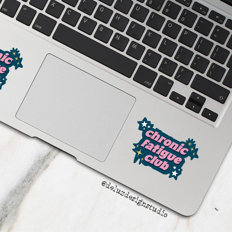 May include: A laptop with a keyboard and trackpad, featuring a sticker that reads "Chronic Fatigue Club" in pink and teal, surrounded by stars and floral accents. The sticker is on the trackpad.