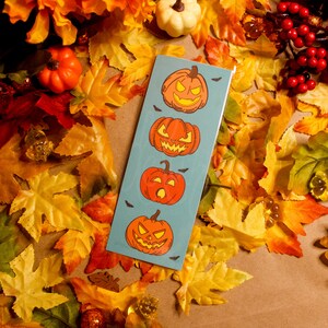 Könnte beinhalten: Ein blaues Lesezeichen mit fünf orangefarbenen Kürbisgesichtern. Das Lesezeichen ist von Herbstblättern umgeben.