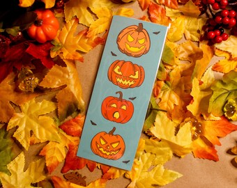 Könnte beinhalten: Ein blaues Lesezeichen mit fünf orangefarbenen Kürbisgesichtern. Das Lesezeichen ist von Herbstblättern umgeben.