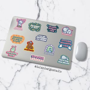 K&ouml;nnte beinhalten: Ein Laptop, der mit bunten Aufklebern mit verschiedenen Designs und Texten bedeckt ist. Aufkleber enthalten S&auml;tze wie "chronic fatigue club", "invisible disability club" und "flare day, dnd please". Eine Computermaus ist rechts zu sehen.