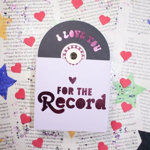 May include: A purple and white card with a black vinyl record attached to it. The card says "For the Record" in a pink and purple font. The vinyl record has a pink and white heart on it and says "I Love You" in pink and white letters.