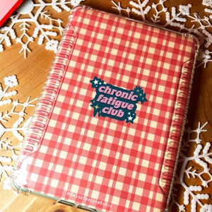 May include: A clear, protective case with a red and cream gingham pattern. The case features the text "chronic fatigue club" in a colorful sticker design. The case is designed to fit a tablet or notebook.