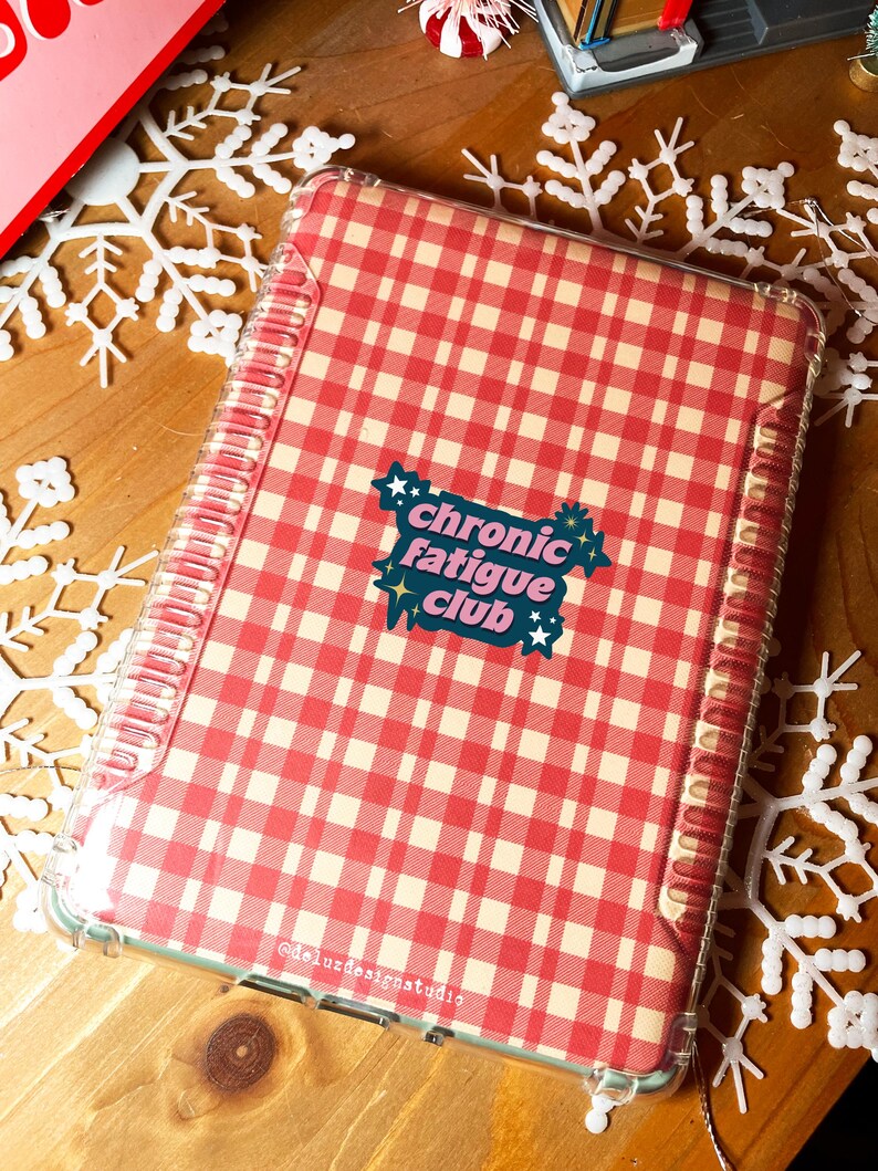 May include: A clear, protective case with a red and cream gingham pattern. The case features the text "chronic fatigue club" in a colorful sticker design. The case is designed to fit a tablet or notebook.