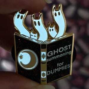 Könnte beinhalten: Ein goldfarbener Emaille-Pin mit einem schwarzen Buch mit dem Text "GHOST summoning for DUMMIES". Der Pin hat weiße Geisterfiguren und ein Mond-Design. Der Pin wird von einer Person gehalten.