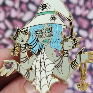 May include: A gold-colored enamel pin featuring a witch with blue hair, glasses, and a white hat adorned with spiders. The witch is holding spider-themed accessories. The pin has a white spiderweb design on the witch's dress.