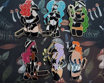 May include: Set of seven enamel pins featuring women wearing black outfits and plague doctor masks. Each pin has a different color hair and a unique accessory, such as a chain, a saw, or a whip.