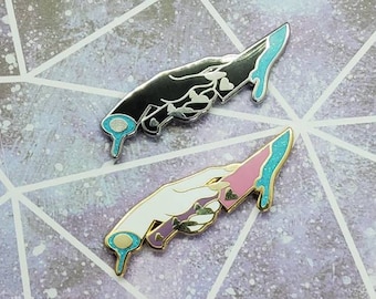 May include: Two enamel pins depicting hands holding dripping knives. One pin features a black hand with a silver knife and blue dripping paint. The other pin features a white hand with a gold knife and pink dripping paint.