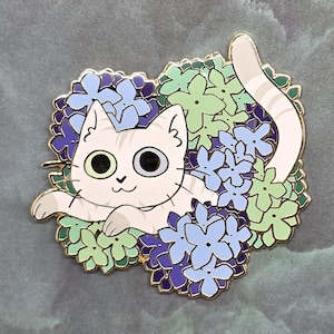May include: A gold-rimmed enamel pin featuring a light cream-colored cat with faint stripes resting among clusters of blue, light blue, and pale green hydrangea flowers. The cat has large, dark eyes and a small nose, with its tail curled upward.