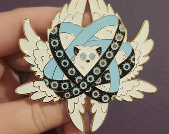 May include: A gold-tone enamel pin featuring a white furry creature with large eyes, surrounded by a blue and black ribbon with white polka dots. The creature has wings and is surrounded by a halo of white glitter.