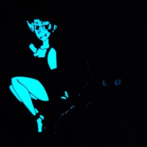 May include: A blue and black glow-in-the-dark pin featuring a woman with pointed ears and a cat with glowing eyes.