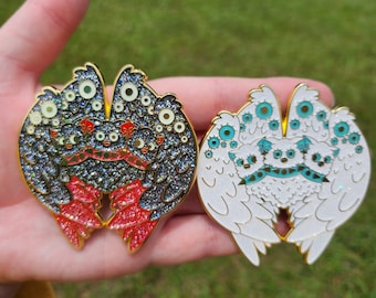 May include: Two enamel pins depicting a creature with multiple eyes and wings. The first pin is black with red accents and glitter. The second pin is white with blue accents and glitter.