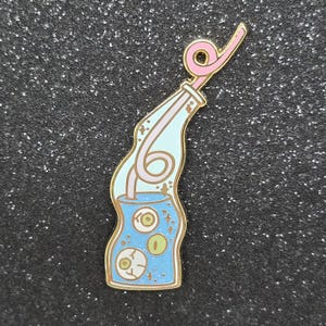Könnte beinhalten: Eine Metall-Emaille-Anstecknadel in Form einer gewellten Glasflasche, gefüllt mit blauer Flüssigkeit und drei schwebenden Augäpfeln. Ein rosa, geschwungener Strohhalm ragt aus der Flaschenöffnung. Das Design hat goldene Umrandungen und kleine Sterne.