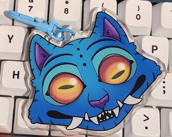 May include: A blue cat-shaped keychain with a cartoon design. The cat has large yellow eyes, purple accents, and sharp teeth. It includes a blue clasp for attachment. The keychain is resting on a keyboard.
