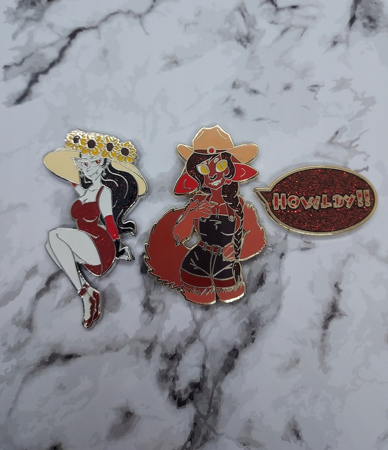 May include: Three enamel pins featuring cartoon characters. The first pin is a woman with long black hair wearing a red dress and a sunflower crown. The second pin is a red-furred character wearing a cowboy hat and overalls. The third pin is a speech bubble with the text "HOWLDY!!" in red glitter.