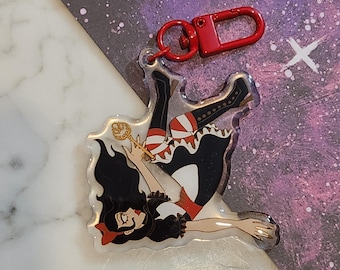 May include: A clear acrylic keychain featuring a cartoon illustration of a woman with long black hair, wearing a red and white dress, falling backwards while holding a golden key. The keychain has a red metal ring.