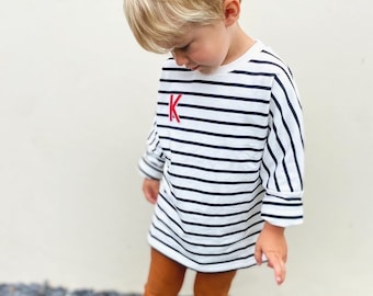 May include: A child wearing a white and black striped long-sleeved shirt with a red "K" on the front. The child is also wearing brown leggings and is standing on a gray surface.
