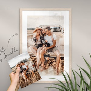 May include: A family of three, a man, a woman, and a young girl, are sitting in the back of a pickup truck. They are smiling and looking at the camera. The truck is parked in a field of tall grass. The image is in a light brown frame.