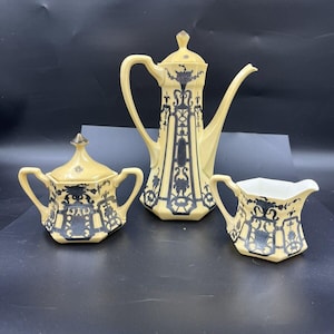May include: A vintage tea set with a cream-colored base and dark blue decorative accents. The set includes a coffee pot, a sugar bowl with a lid, and a creamer. The design features ornate patterns and a classic aesthetic.