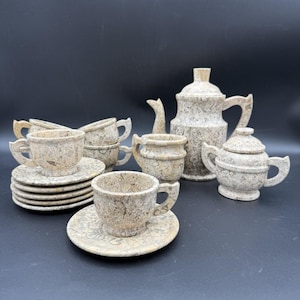 May include: A complete tea set with a speckled, stone-like appearance. The set includes a teapot, sugar bowl, creamer, cups, and saucers. The cups and saucers are stacked neatly, and the teapot has a lid.