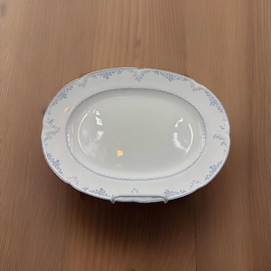 May include: An oval, white serving platter with a delicate blue floral pattern around the rim. The platter is supported by a silver metal stand, and is set against a light brown wooden surface. The design is classic and elegant.