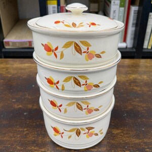May include: A stack of four white ceramic bowls with lids, featuring a floral design in orange, yellow, and pink. The bowls have a gold trim and are stacked on top of each other. The top bowl has a lid with a small knob.