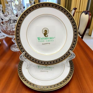 May include: Two white Waterford Fine China plates with gold rims and black and gold patterned borders. Each plate has the Waterford logo and a seahorse emblem. The plates are displayed on a clear stand.
