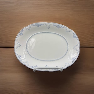 May include: White oval serving platter with a scalloped edge and blue floral design. The platter has a blue border and a decorative rim. The serving dish is displayed on a white metal stand.