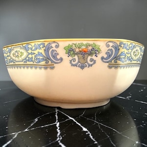 May include: Cream-colored ceramic bowl with a decorative fruit basket and scroll design. The bowl has a gold rim and blue and yellow accents. The fruit basket includes grapes, apples, and other fruits. The bowl is sitting on a black surface.