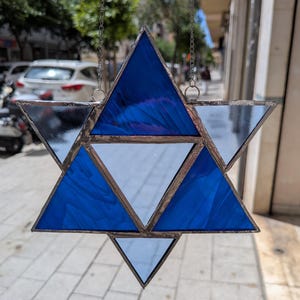 Puede incluir: Un atrapasoles de vidrio manchado con forma de Estrella de David. La estrella de seis puntas presenta triángulos de diferentes tonos de azul y vidrio transparente, enmarcados con metal oscuro. El atrapasoles está suspendido por una cadena.