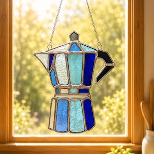 Puede incluir: Un atrapasoles de vidrieras con forma de cafetera moka, que presenta varios paneles de vidrio en tonos azul, azul claro y blanco unidos por líneas de soldadura de metal plateado, suspendido por una fina cadena plateada.