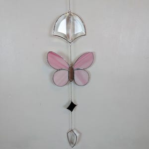 Puede incluir: Adorno colgante de vidrieras. Presenta una mariposa con alas rosas y cuerpo marrón, una forma de paraguas transparente, un diamante negro y una lágrima transparente. Todas las piezas están unidas por una cadena plateada, creando un adorno colgante.