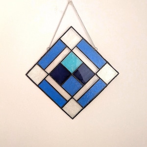 Puede incluir: Un panel de vidrieras con un diseño geométrico. El panel presenta una forma de diamante con un borde negro y está compuesto por piezas de vidrio azul, blanco y turquesa. Se adjunta una cadena plateada para colgarlo.
