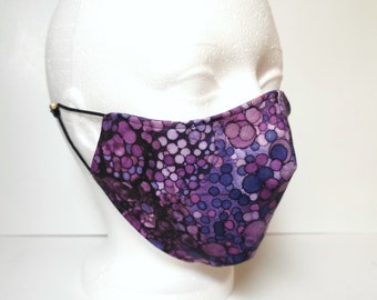 May include: A fabric face mask with a pattern of purple and blue circles. The mask is on a white mannequin head. The mask has black elastic ear loops with gold-colored adjusters.