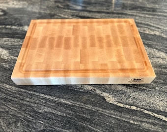 May include: A light-colored, rectangular end-grain butcher block cutting board. The board has a juice groove around the perimeter and a logo on the corner. The wood grain pattern is visible, and the board is made of multiple wood pieces.