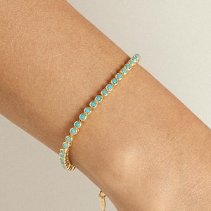 May include: Gold-toned bracelet featuring a row of small, round, turquoise-colored gemstones. The bracelet is worn on a person's arm, showcasing the delicate design and vibrant color of the stones. The clasp is visible at the end of the bracelet.
