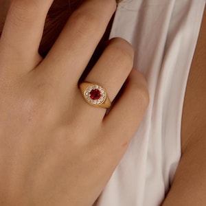 May include: Gold ring featuring a round, red gemstone center, surrounded by a halo of smaller, clear stones. The ring is set on a gold band. The ring is worn on a finger.
