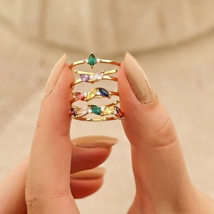 May include: A set of five gold rings with colorful marquise-shaped gemstones. The rings are stacked on top of each other and are being held by a hand.