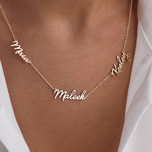 May include: A gold-colored necklace with three name pendants: "Maia", "Maleek", and "Keeley" in a delicate script font. The necklace is displayed against a white shirt.