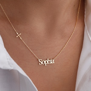 May include: A gold necklace with a delicate chain and a small cross charm. The necklace also features a nameplate pendant with the name "Sophia" in a simple, elegant font.