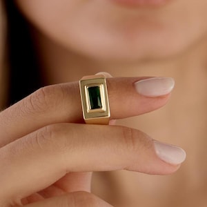 May include: A gold-colored ring with a rectangular, dark green stone. The ring is held up by a person's fingers, showcasing the jewelry's design and color. The ring's band is thick and rectangular, with a polished finish.
