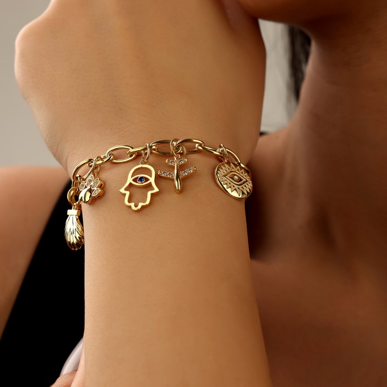 May include: Gold charm bracelet featuring a variety of charms, including a hamsa hand, an evil eye, a dragonfly, and a seashell. The charms are attached to a gold chain.