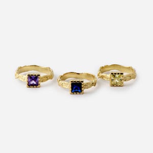 May include: Three gold rings with textured bands and square-shaped gemstones. The rings feature a purple, blue, and yellow gemstone.