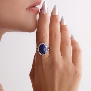 May include: Silver ring with an oval, deep blue gemstone. The ring has a twisted rope design around the gemstone. The ring is on a hand with long, polished nails.