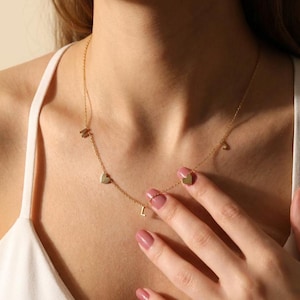 May include: A delicate gold necklace with heart-shaped charms and small letter pendants. The necklace is worn around the neck, with the charms and letters resting on the chest. The letters and hearts are gold in color.