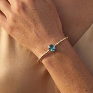 May include: A gold-colored bracelet with a twisted rope design. The bracelet features a bright blue, oval-shaped gemstone. The jewelry is worn on a wrist.