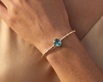 Könnte beinhalten: Ein goldfarbenes Armband mit einem gedrehten Seildesign. Das Armband hat einen hellblauen, oval geformten Edelstein. Der Schmuck wird am Handgelenk getragen.