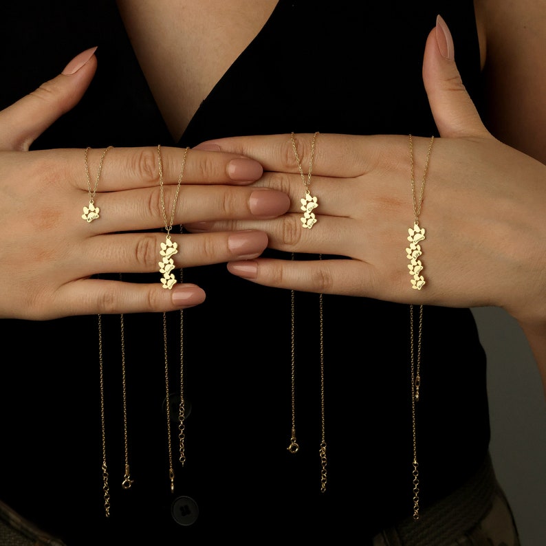 May include: Four gold necklaces with delicate paw print charms. The necklaces are layered and feature different lengths.