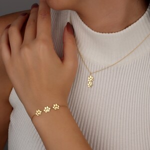 May include: A gold necklace and bracelet with paw print charms. The necklace has two charms and the bracelet has three charms. Each charm has a name engraved on it.