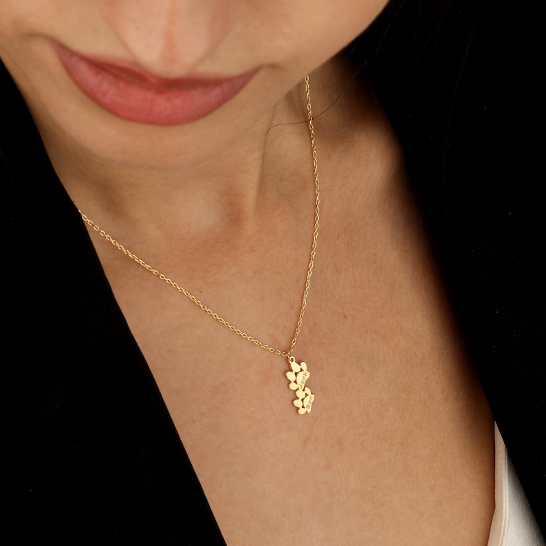 May include: A gold chain necklace with a pendant featuring a cluster of paw prints. The paw prints are arranged in a vertical line and have small engraved letters on them.