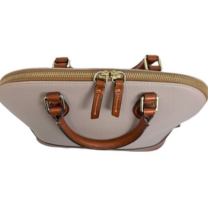 May include: A light gray dome-shaped handbag with brown leather handles and accents. The bag has a gold zipper and two brown leather zipper pulls. The handles are attached with gold-colored hardware.